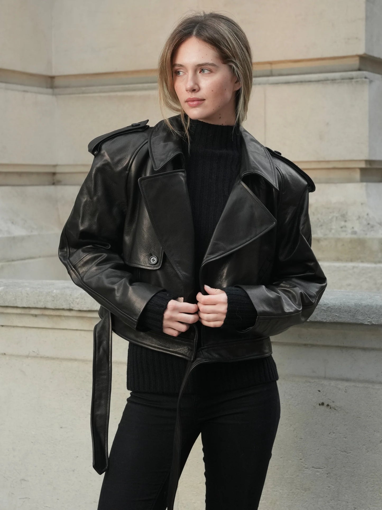 Woman wearing a black leather jacket and black outfit standing against a stone wall.
