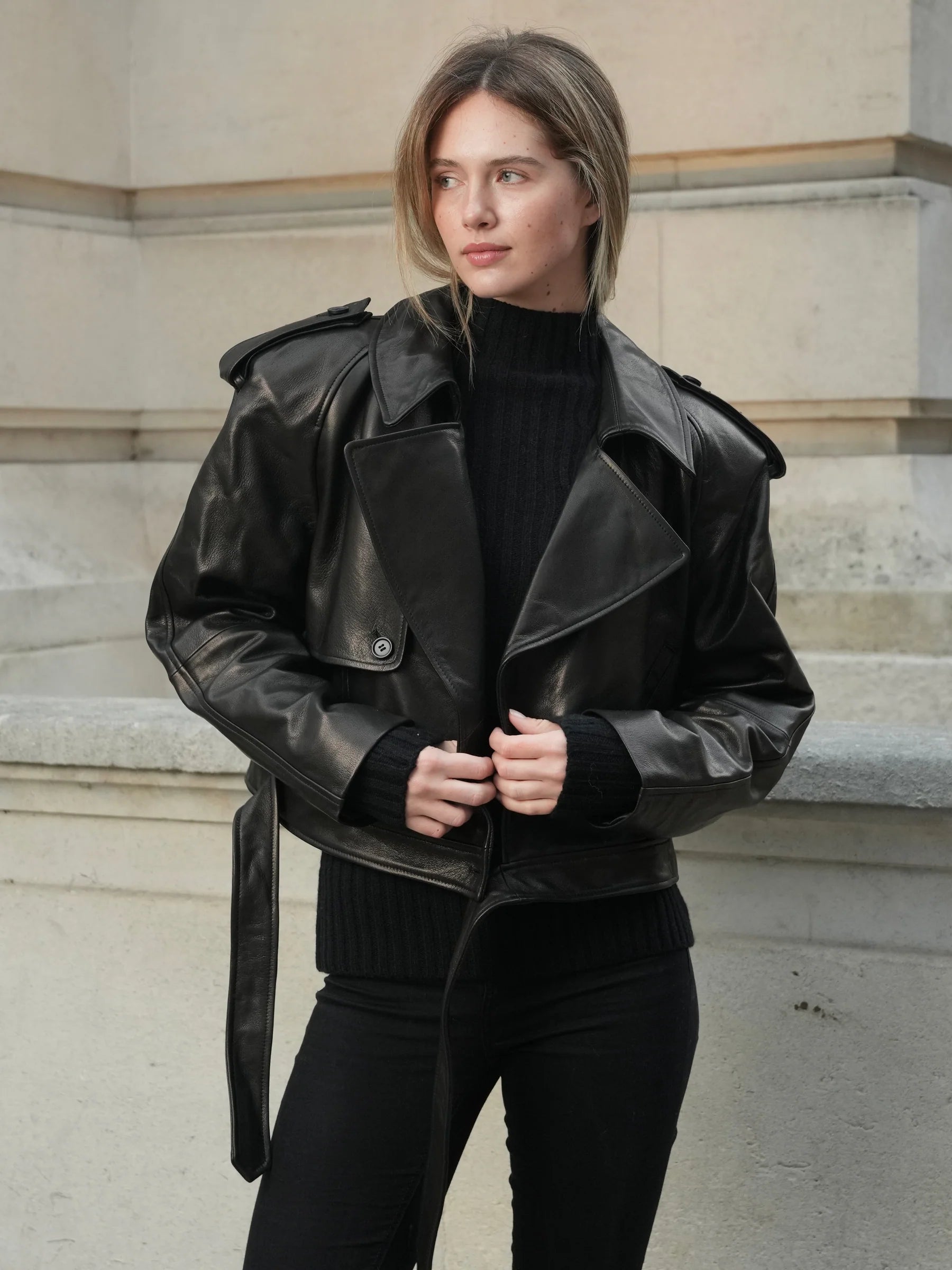 Woman wearing a black leather jacket and black outfit standing against a stone wall.